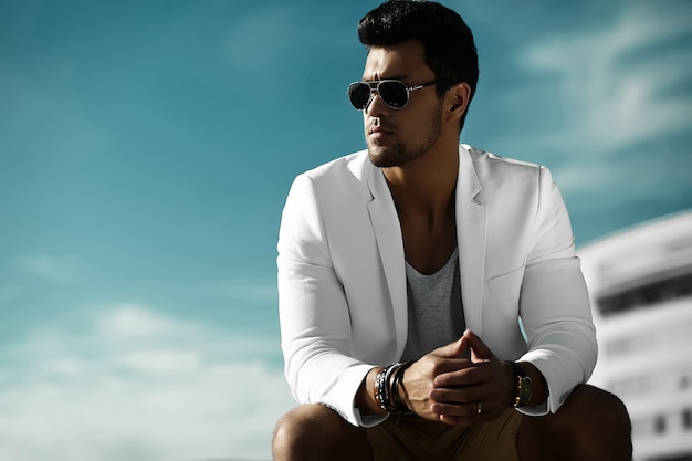Foto gratuita retrato de moda de hombre de negocios guapo joven modelo guapo hombre en traje casual de tela en gafas de sol sentado en la calle detrás del cielo azul