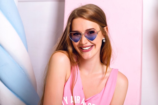 Foto gratuita retrato de moda interior de moda de increíble hermosa joven posando cerca de grandes macarrones falsos y dulces. feliz chica rubia con gafas de sol de corazones mano cerca de la cara. sonrisa satisfecha