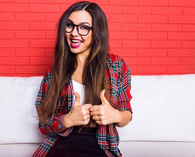 Foto gratuita retrato de moda interior de mujer joven bonita hipster divirtiéndose y sonriendo, vistiendo ropa casual. muro urbano rojo brillante.