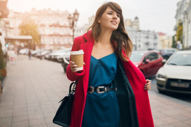 Retrato de moda de joven hermosa mujer elegante caminando en la calle de la ciudad en abrigo rojo, tendencia de estilo otoñal, tomando café, sonriente, feliz, con vestido de seda azul