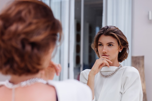 Retrato de moda de joven mujer caucásica modelo profesional en blazer blanco y cadena de plata mirar en espejo en villa de lujo