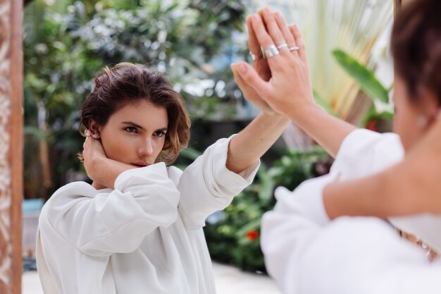 Retrato de moda de joven mujer caucásica modelo profesional en blazer blanco y cadena de plata mirar en espejo en villa de lujo