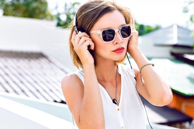 Retrato de moda de joven mujer sexy escuchando su música favorita en sus auriculares, maquillaje brillante, colores frescos de verano de cerca. Posando en el techo, estado de ánimo positivo.