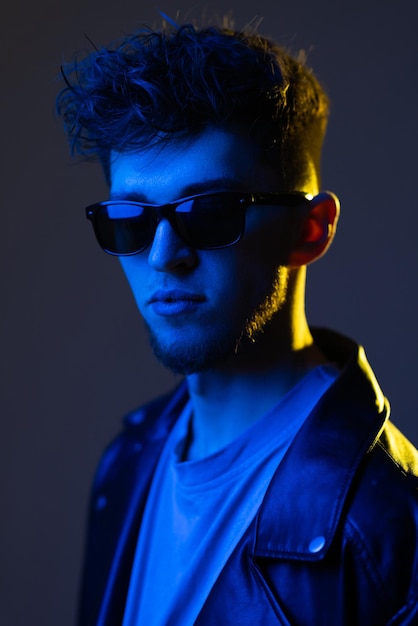 Retrato de moda masculina de un chico afroamericano guapo y confiado con elegante chaqueta de cuero y gafas de sol posando bajo luces de neón sobre fondo de estudio púrpura