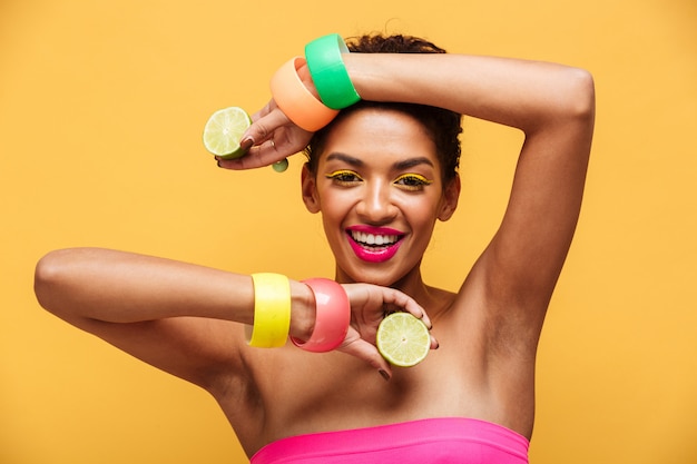 Retrato de moda de mujer afroamericana con accesorios de moda posando en la cámara con dos partes de limón maduro fresco en manos aisladas, sobre pared amarilla