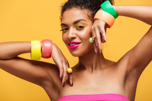 Retrato de moda de mujer afroamericana atractiva con maquillaje brillante mostrando joyas en sus manos aisladas, sobre pared amarilla