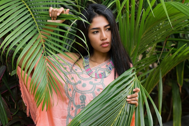 Retrato de moda de mujer asiática atractiva posando en jardín tropical. Lleva un vestido boho y accesorios elegantes.