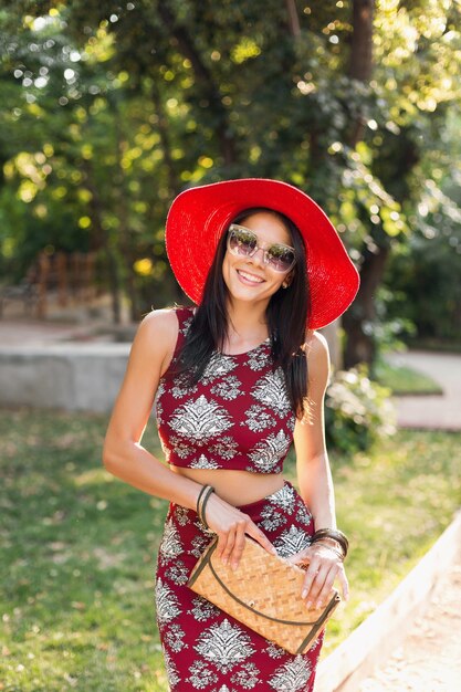 Retrato de moda de mujer elegante atractiva sonriente caminando en el parque en traje de verano vestido estampado, con accesorios de moda, bolso, gafas de sol, sombrero rojo, relajarse en vacaciones