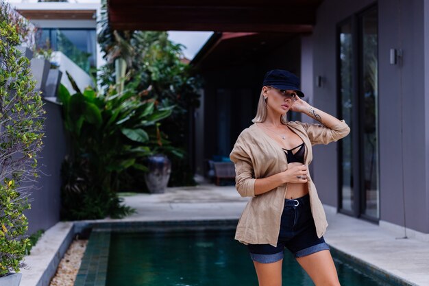 Retrato de moda de mujer elegante caucásica rubia en chaqueta de gorra francesa y pantalones cortos al aire libre fuera de la villa