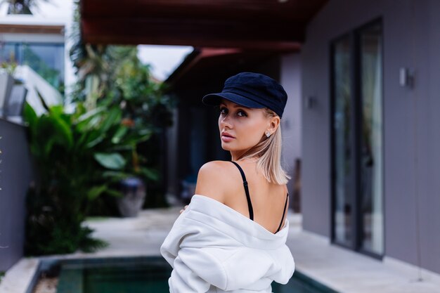 Retrato de moda de mujer elegante caucásica rubia en chaqueta de gorra francesa y pantalones cortos al aire libre fuera de la villa