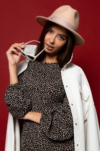 Retrato de moda de mujer elegante morena con bata blanca vestida y sombrero beige con gafas de sol. estilo de moda de otoño o invierno.