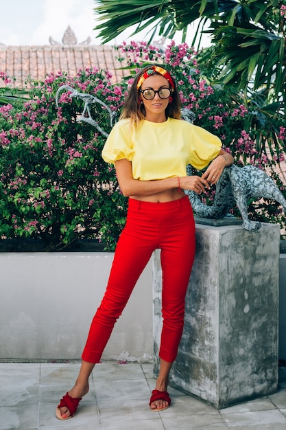 Retrato de moda de mujer elegante en pantalones rojos, top amarillo, aretes, gafas de sol y pañuelo en la cabeza.