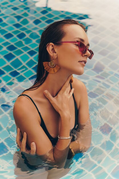 Retrato de moda de mujer elegante seductora en elegantes aretes amarillos con cuerpo perfecto posando en la piscina durante las vacaciones en resort de lujo.