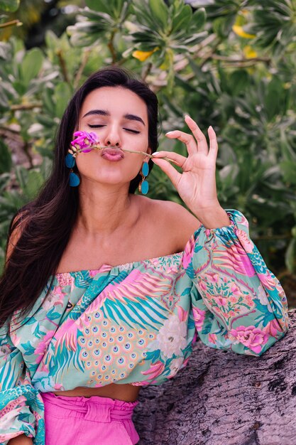 Retrato de moda de mujer elegante en top de manga larga con estampado de colores y pantalones cortos de color rosa en la playa, fondo tropical.