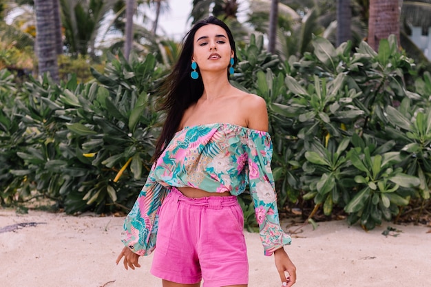 Foto gratuita retrato de moda de mujer elegante en top de manga larga con estampado de colores y pantalones cortos de color rosa en la playa, fondo tropical.