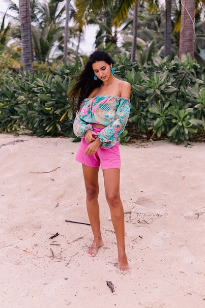 Retrato de moda de mujer elegante en top de manga larga con estampado de colores y pantalones cortos de color rosa en la playa, fondo tropical.