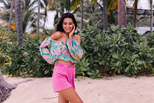 Retrato de moda de mujer elegante en top de manga larga con estampado de colores y pantalones cortos de color rosa en la playa, fondo tropical.