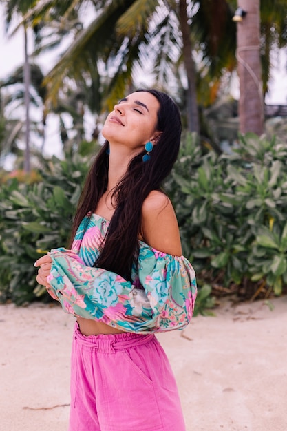 Retrato de moda de mujer elegante en top de manga larga con estampado de colores y pantalones cortos de color rosa en la playa, fondo tropical.