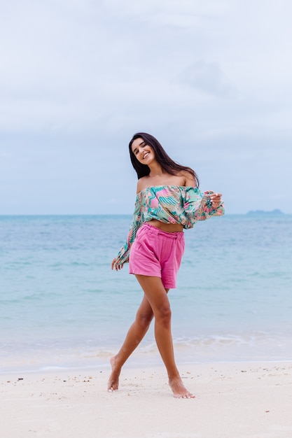 Retrato de moda de mujer elegante en top de manga larga con estampado de colores y pantalones cortos de color rosa en la playa, fondo tropical.