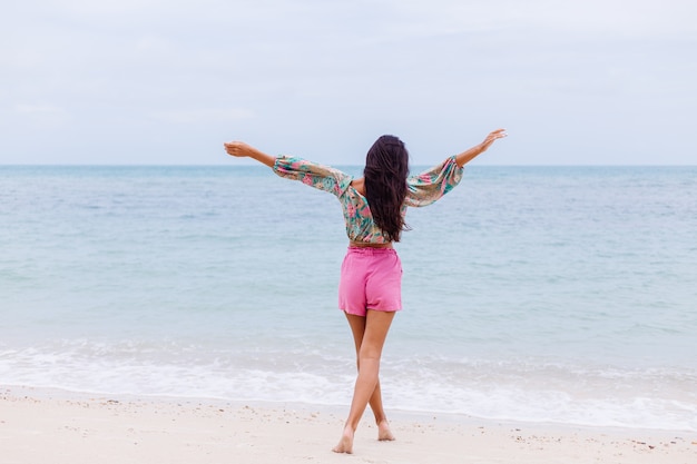 Retrato de moda de mujer elegante en top de manga larga con estampado de colores y pantalones cortos de color rosa en la playa, fondo tropical.