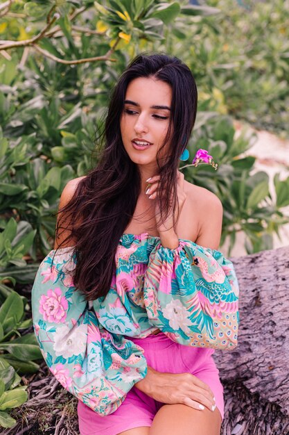 Retrato de moda de mujer elegante en top de manga larga con estampado de colores y pantalones cortos de color rosa en la playa, fondo tropical.