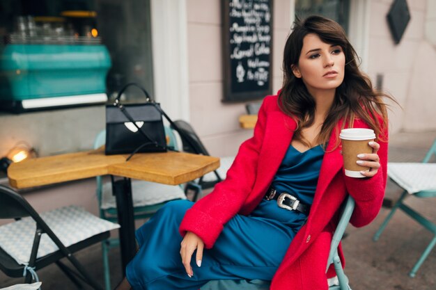 Retrato de moda de mujer joven y atractiva con estilo sentado en el café de la calle de la ciudad en abrigo rojo, tendencia de estilo otoñal, tomando café, vestido de seda azul, dama elegante, mirando hacia adelante