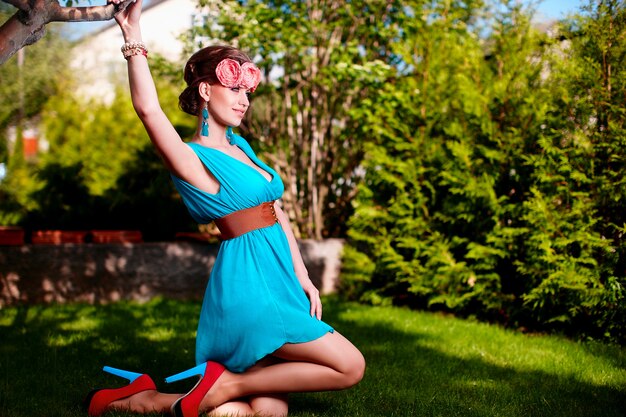 Retrato de moda de mujer joven y bella modelo dama mujer con peinado en vestido azul brillante posando al aire libre sentado en la hierba verde cerca de bush con flores en el cabello