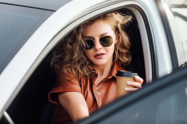 Retrato de moda de mujer joven en coche blanco con taza o café