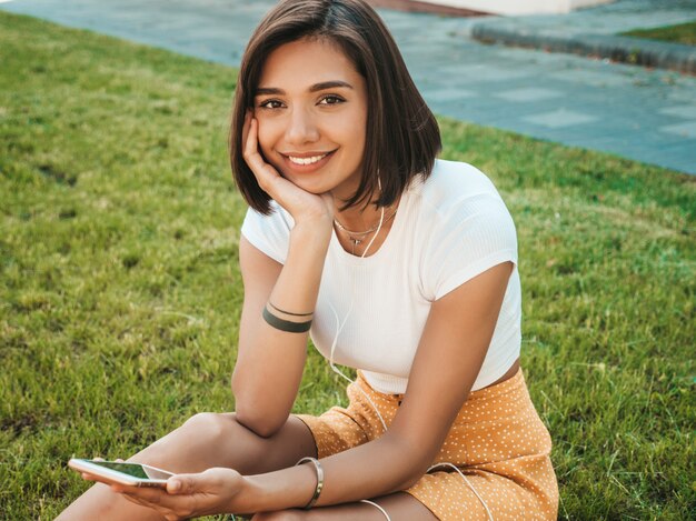 Retrato de moda de mujer joven con estilo hipster. Chica con lindo traje de moda. Modelo sonriente disfrutar de sus fines de semana, sentada en el parque. Mujer escuchando música a través de auriculares