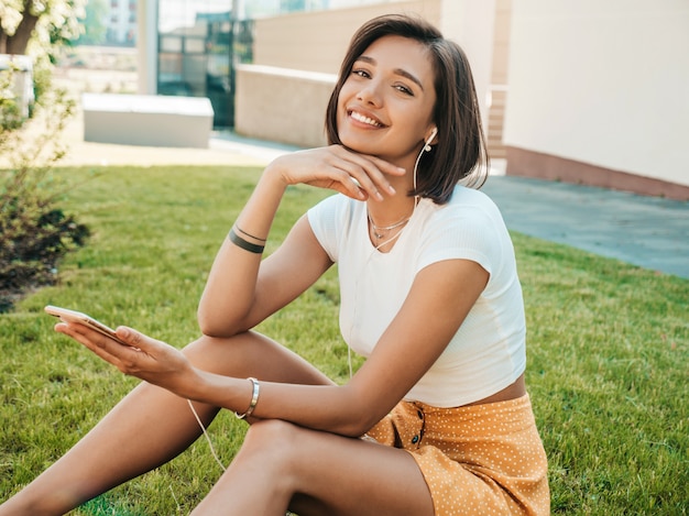 Retrato de moda de mujer joven con estilo hipster. Chica con lindo traje de moda. Modelo sonriente disfrutar de sus fines de semana, sentada en el parque. Mujer escuchando música a través de auriculares