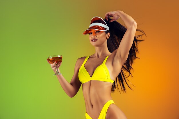 Retrato de moda de mujer joven en forma y deportiva con cóctel en elegante traje de baño de lujo amarillo en pared degradada cuerpo perfecto listo para el verano