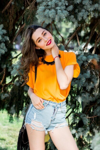Retrato de moda de una mujer joven y guapa en el parque de verano con camisa naranja y elegantes pantalones cortos de jeans con bolso boho