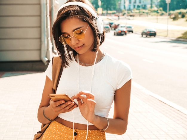 Retrato de moda de mujer joven inconformista elegante caminando en la calle. Chica con lindo traje de moda. Modelo sonriente disfrutar de sus fines de semana, viajar con mochila. Mujer escuchando música a través de auriculares