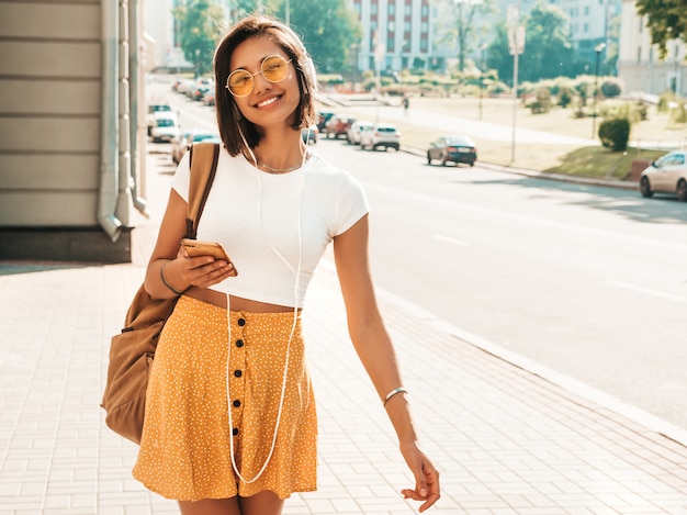 Foto gratuita retrato de moda de mujer joven inconformista elegante caminando en la calle. chica con lindo traje de moda. modelo sonriente disfrutar de sus fines de semana, viajar con mochila. mujer escuchando música a través de auriculares