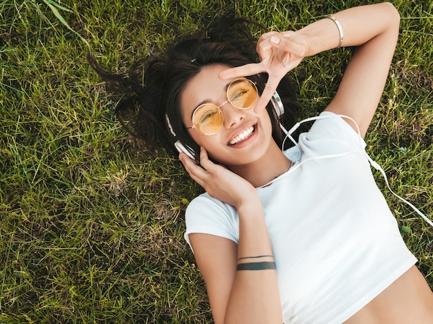 Retrato de moda de mujer joven inconformista elegante tirado en el pasto en el parque. Chica teje atuendo de moda. Modelo sonriente disfrutar de sus fines de semana. Mujer escuchando música a través de auriculares. Vista superior