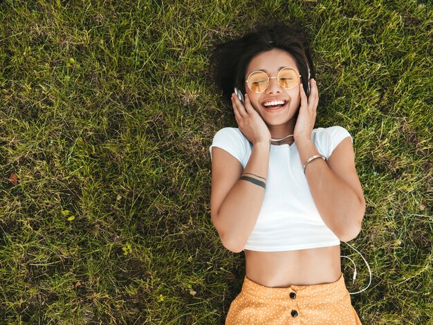 Retrato de moda de mujer joven inconformista elegante tirado en el pasto en el parque. Chica teje atuendo de moda. Modelo sonriente disfrutar de sus fines de semana. Mujer escuchando música a través de auriculares. Vista superior