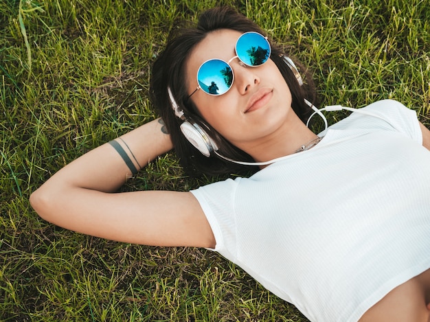 Retrato de moda de mujer joven inconformista elegante tirado en el pasto en el parque. Chica teje atuendo de moda. Modelo sonriente disfrutar de sus fines de semana. Mujer escuchando música a través de auriculares. Vista superior