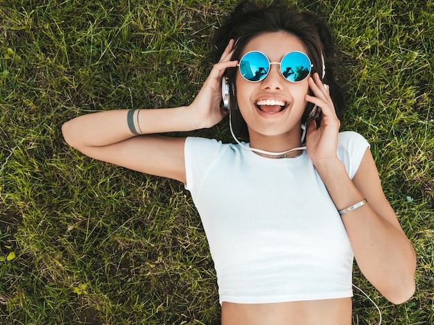 Retrato de moda de mujer joven inconformista elegante tirado en el pasto en el parque. Chica teje atuendo de moda. Modelo sonriente disfrutar de sus fines de semana. Mujer escuchando música a través de auriculares. Vista superior