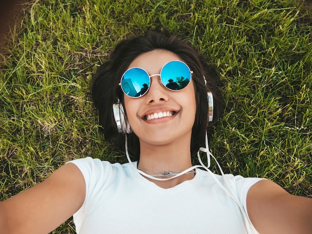 Retrato de moda de mujer joven inconformista elegante tirado en el pasto en el parque. Chica teje traje de moda. Modelo sonriente haciendo selfie. Mujer escuchando música a través de auriculares. Vista superior