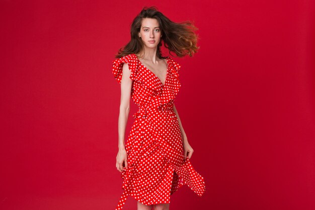 Retrato de moda de mujer joven sonriente atractiva en vestido rojo punteado en estudio rojo