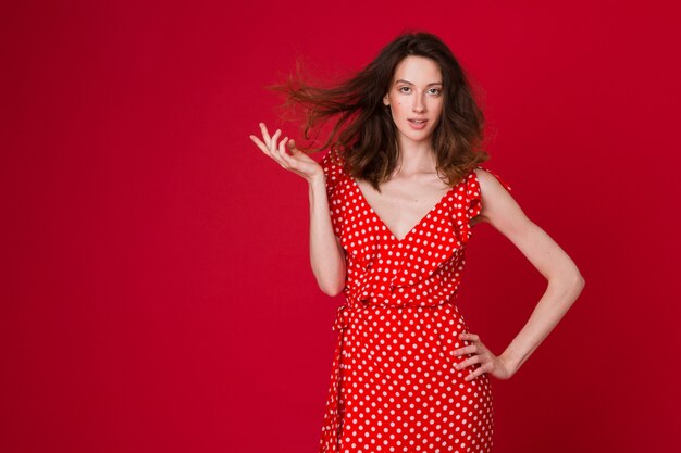 Retrato de moda de mujer joven sonriente atractiva en vestido rojo punteado en estudio rojo