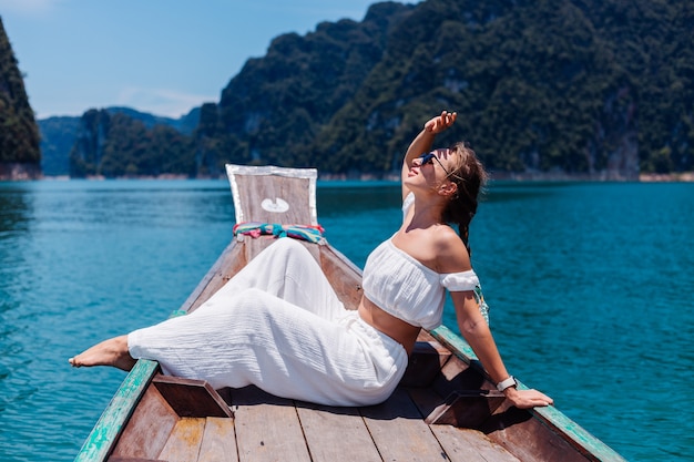 Retrato de moda de mujer joven en top blanco y pantalones de vacaciones, navegando en barco de madera tailandés. Concepto de viaje. Hembra en el parque nacional Khao Sok.