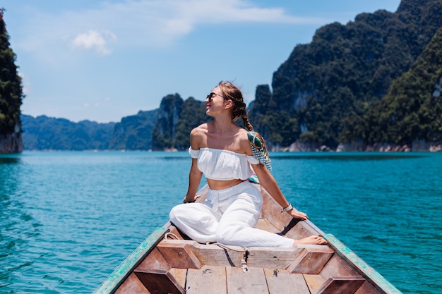 Retrato de moda de mujer joven en top blanco y pantalones de vacaciones, navegando en barco de madera tailandés. Concepto de viaje. Hembra en el parque nacional Khao Sok.