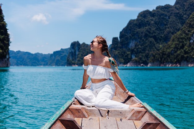 Retrato de moda de mujer joven en top blanco y pantalones de vacaciones, navegando en barco de madera tailandés. Concepto de viaje. Hembra en el parque nacional Khao Sok.