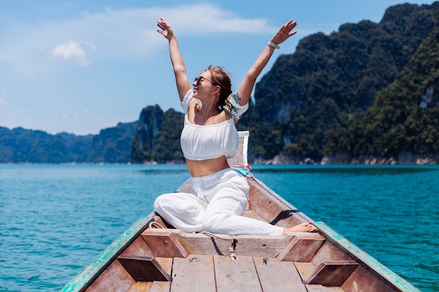 Retrato de moda de mujer joven en top blanco y pantalones de vacaciones, navegando en barco de madera tailandés. Concepto de viaje. Hembra en el parque nacional Khao Sok.