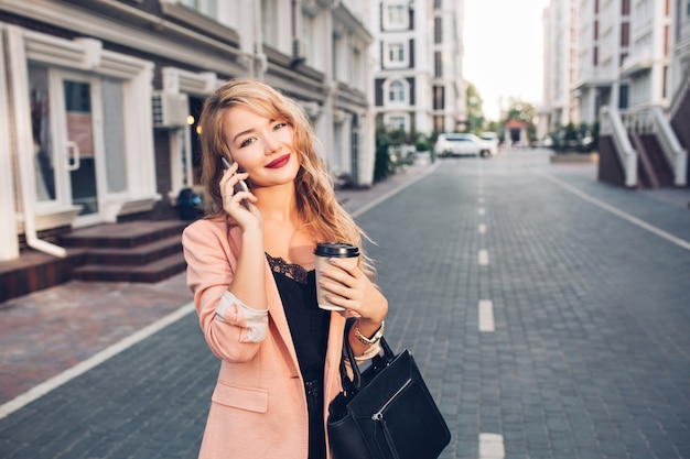 Retrato de moda mujer rubia con pelo largo caminando en chaqueta coral en la calle. Ella está hablando por teléfono, sostiene una taza