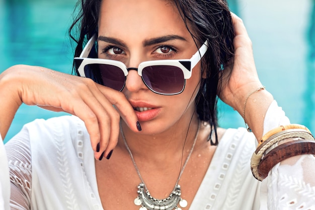 Retrato de moda de mujer sensual increíble con cuerpo bronceado perfecto posando en gafas de sol de moda en la piscina