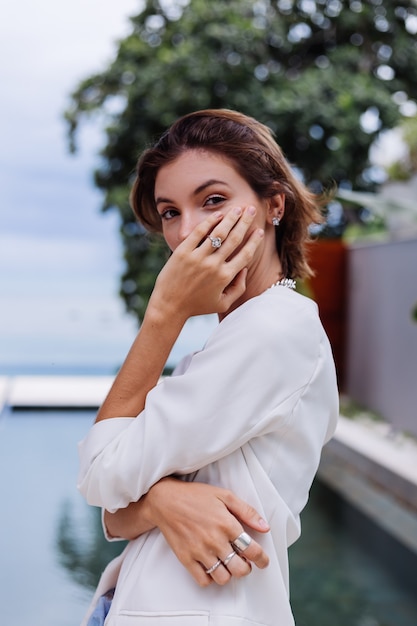 Retrato de moda de mujer en villa de lujo tropical vistiendo elegante blazer blanco y joyas sobre hojas tropicales