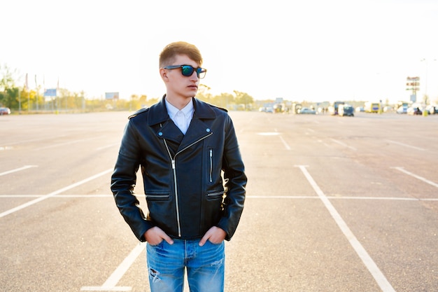 Retrato de moda de otoño al aire libre de hombre elegante posando en el estacionamiento de la ciudad, vestido con gafas de sol y chaqueta de cuero negro de motorista de jeans.