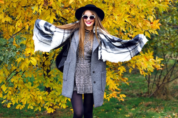 Retrato de moda de otoño de impresionante modelo elegante posando en el parque, hojas doradas y clima fresco, ropa de estilo callejero de lujo, maquillaje brillante, bufanda grande, mini vestido cubre abrigo y sombrero vintage.